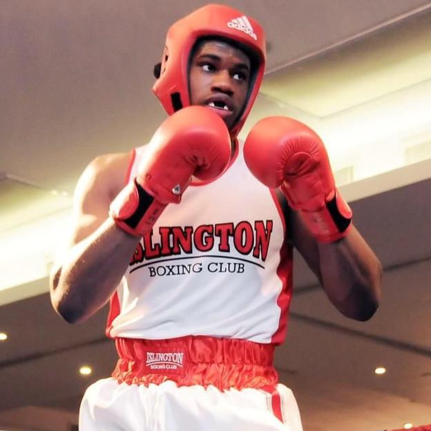 A young boxer in a red headguard, red gloves, and a white and red "ISLINGTON BOXING CLUB" vest.