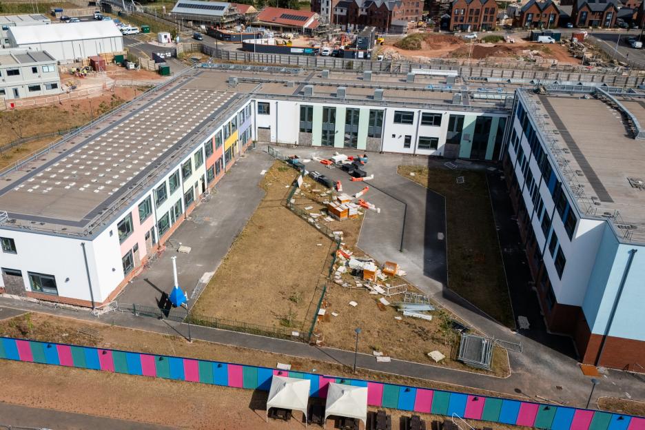Aerial view of the Matford Brook Academy, a large, unused school building with colorful panels on its facade, surrounded by dry grass and construction debris.