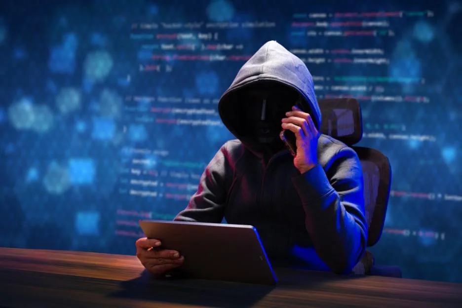 A hooded hacker wearing a mask uses a mobile phone and a tablet against a backdrop of computer code.