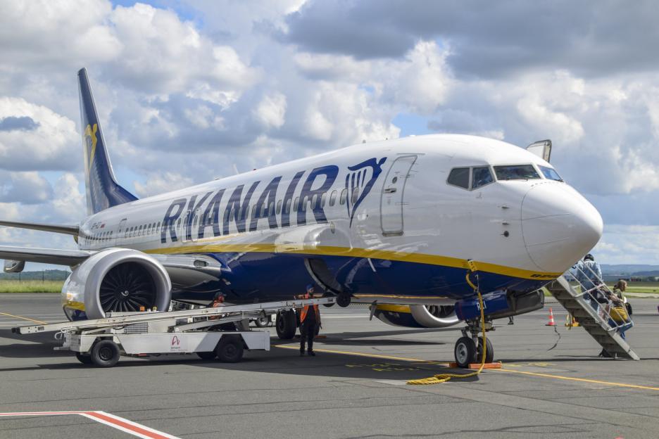 A Ryanair Boeing 737 airplane on the tarmac, with passengers boarding via stairs and ground crew working around the luggage conveyor.