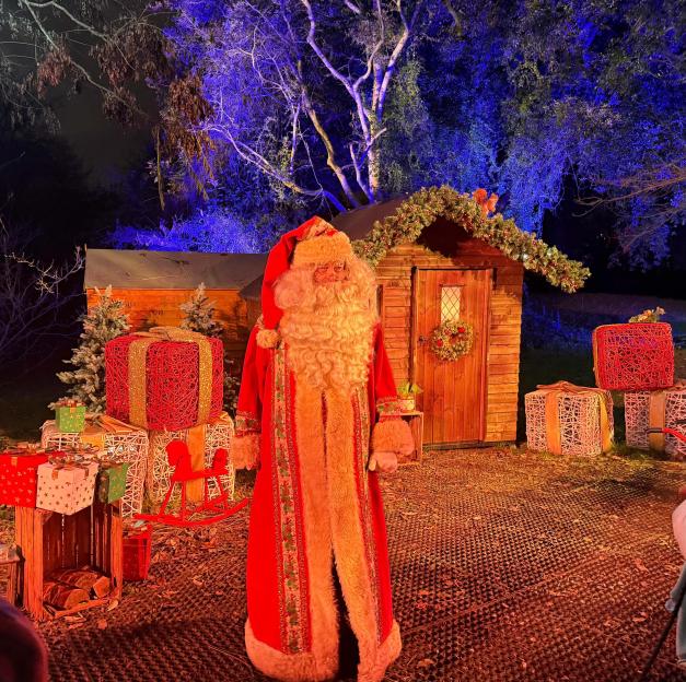 Santa Claus standing in front of a wooden shed decorated with Christmas garlands and surrounded by large, illuminated gift boxes.