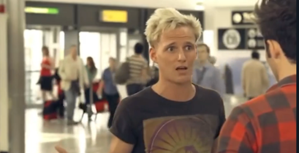 Man with blond hair looks surprised while talking to another man in an airport.