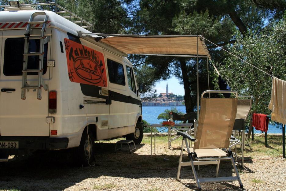 A white camper van with an orange graphic parked under an awning near the sea, with a town visible in the distance.