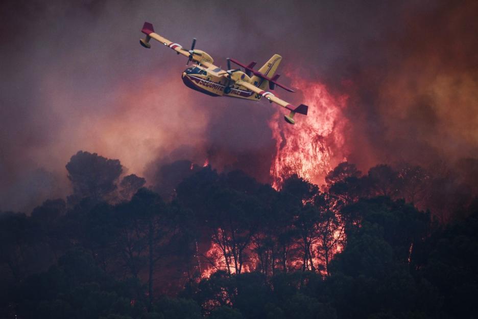 A yellow and red firefighting plane flies over a forest engulfed in flames and smoke.