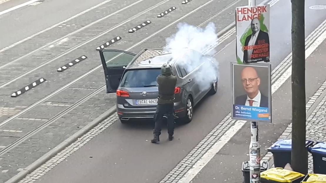 A person shooting a firearm with smoke emanating from the barrel towards a car.
