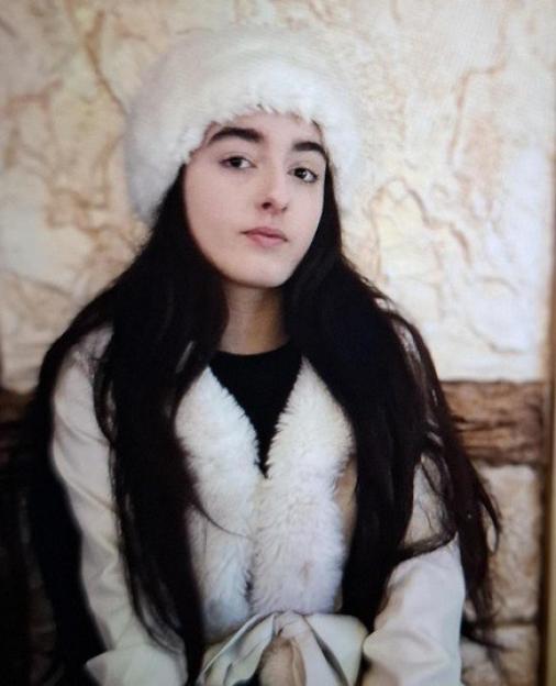 A young woman with long dark hair and a serious expression, wearing a white fur hat and a white coat with a fur collar.