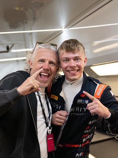 A man with curly white hair and sunglasses on his head, and a young man in a racing suit with "GB4 CHAMPIONSHIP" on it, smile and point at each other.