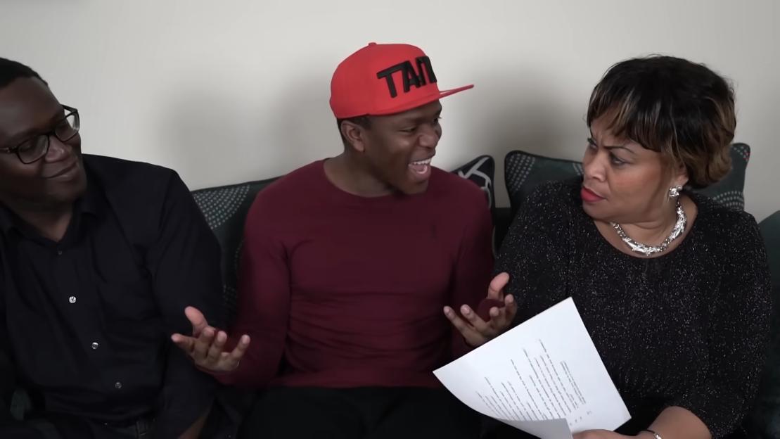 KSI, wearing a red hat and maroon long-sleeved shirt, sits between his parents, talking animatedly and gesturing with his hands as his mother looks on with a surprised expression and holds a sheet of paper, while his father smiles.