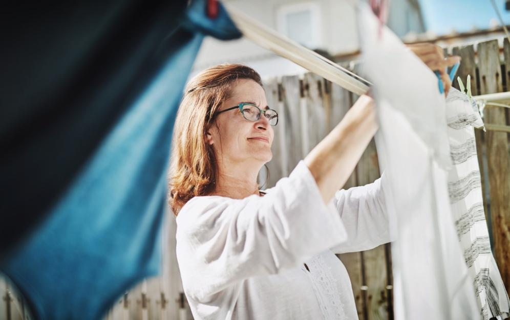 A woman with reddish-brown hair and glasses hangs laundry on a clothesline in her backyard.