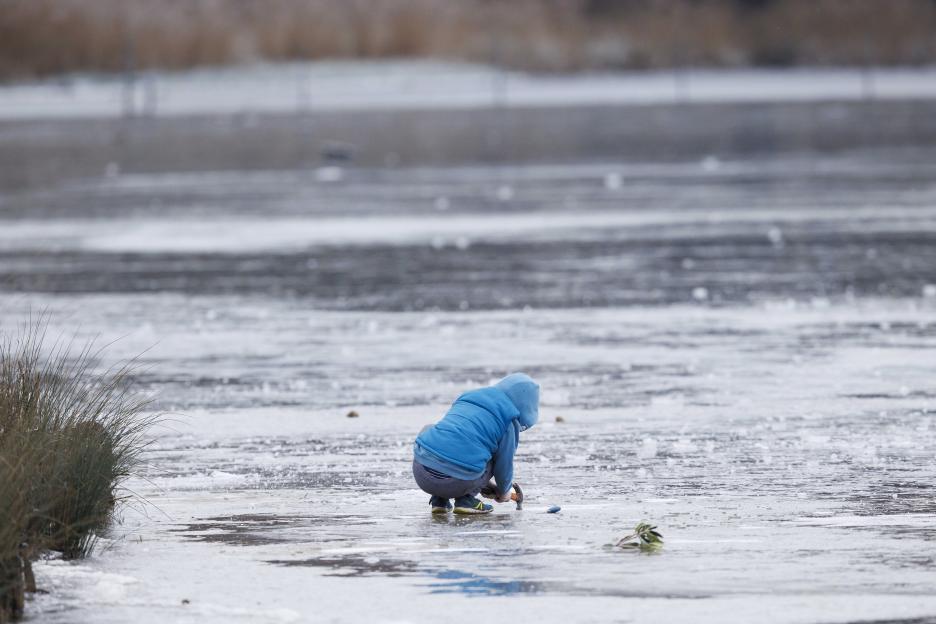 A child in a blue hooded jacket crouches on a frozen pond, hitting the ice with a hammer.
