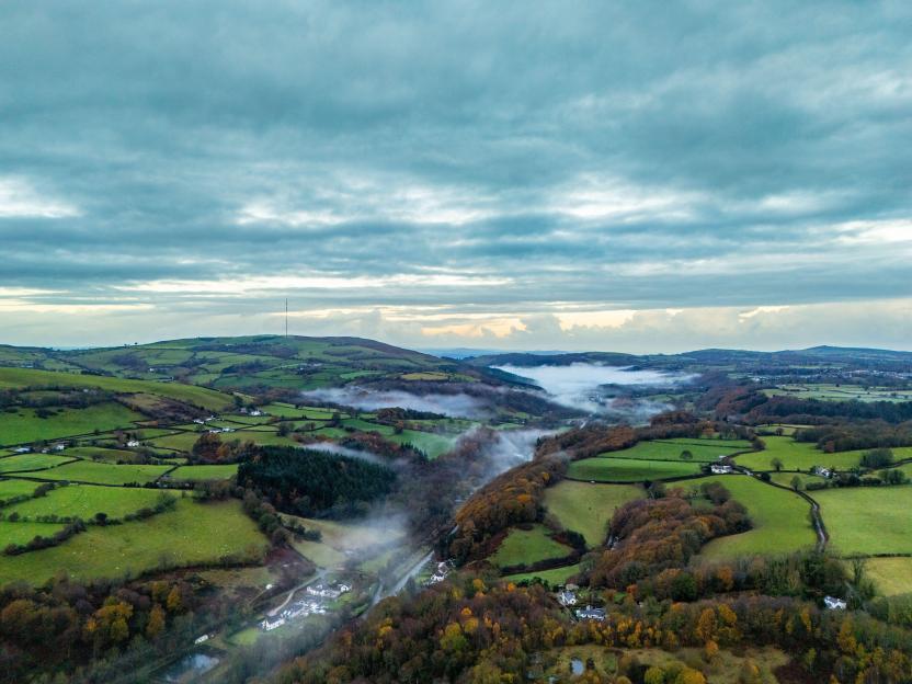 Aerial view of Lixwm village in Flintshire, Wales, with mist in the valley and green fields under a cloudy sky.