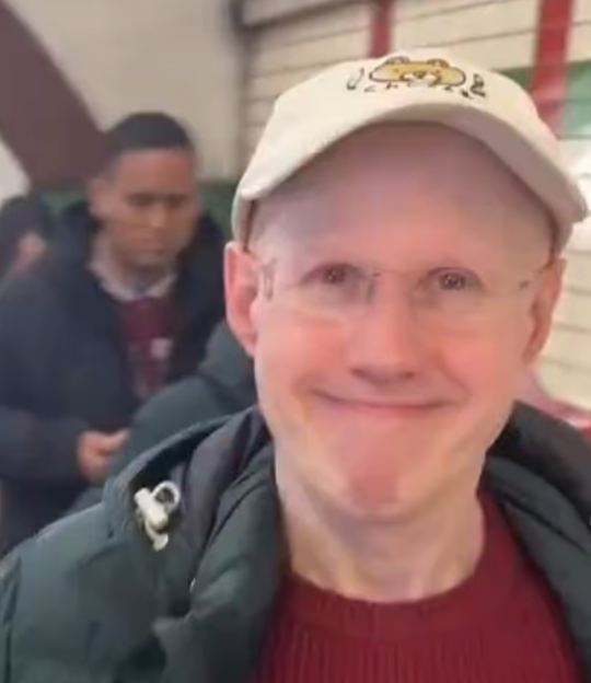 A bald man with glasses and a baseball cap smiles at the camera.