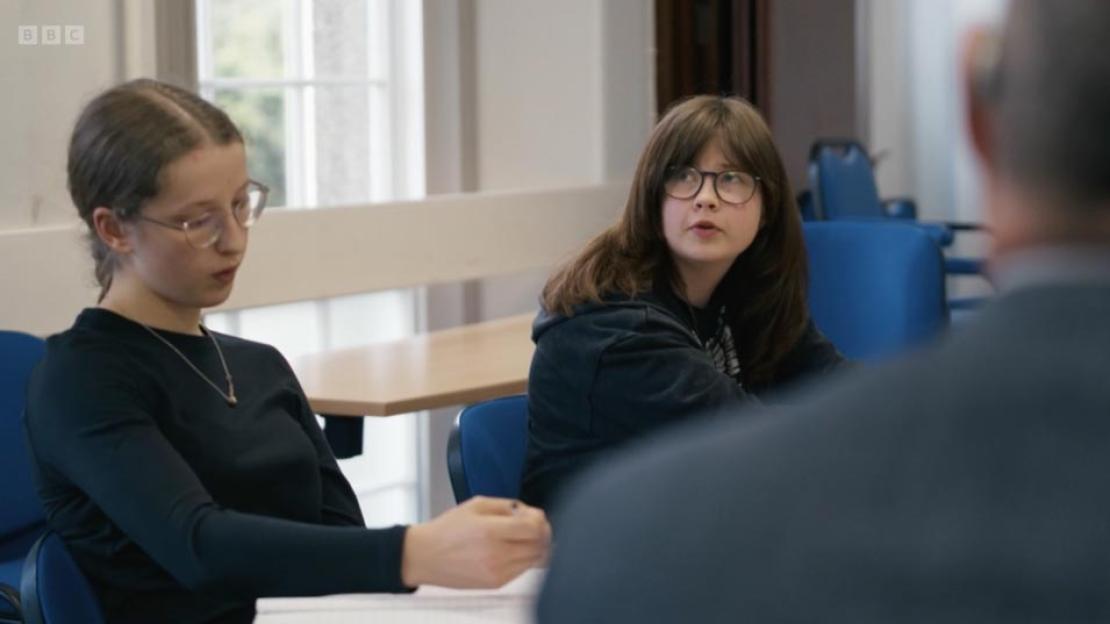 Two young women wearing glasses are seated in a classroom, looking towards an unseen person in the foreground.