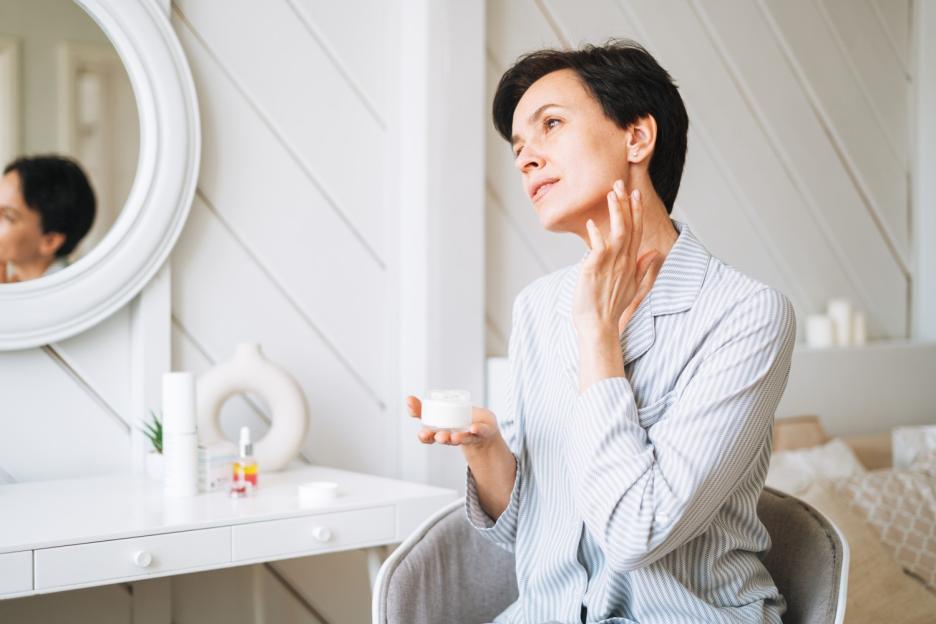 A woman in pajamas applies cream to her neck while sitting at a vanity table.