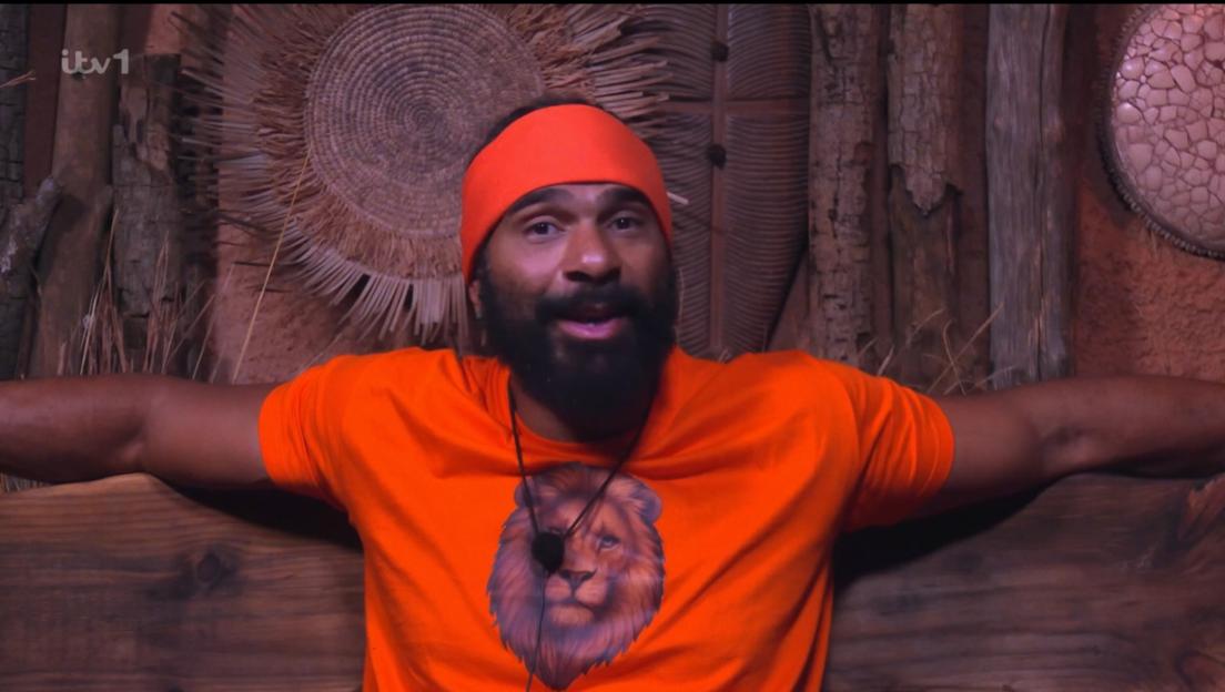 A man wearing an orange bandana and shirt with a lion graphic, smiling with his arms outstretched.