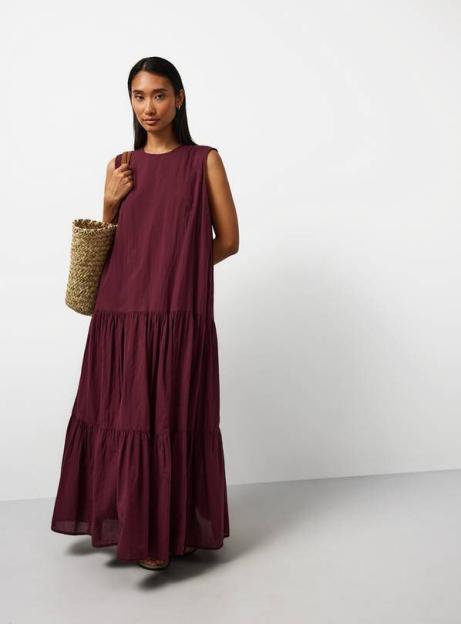 A woman in a long, sleeveless, tiered maroon dress holding a straw bag.