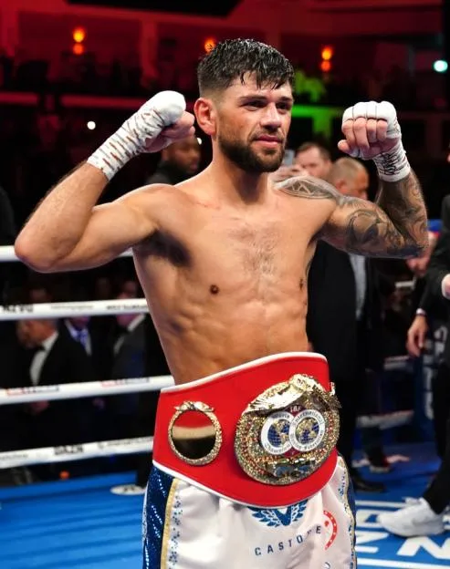 Former boxing world champion Joe Cordina raising his fists in celebration with a championship belt around his waist.
