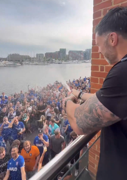 A person spraying champagne onto a crowd of people from a balcony overlooking a marina.