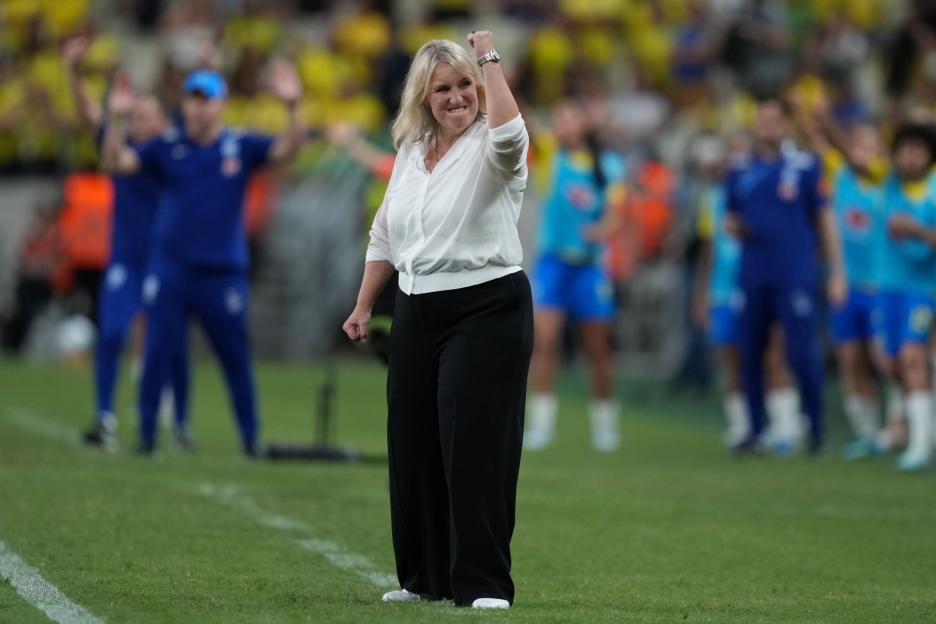 A woman with blonde hair, wearing a white shirt and black pants, pumps her fist in the air on a soccer field.