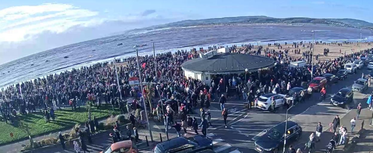 Crowds of people gathered on a beach and street next to the sea with boats in the water at Budleigh Salterton.