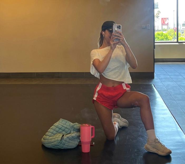 Woman in a cropped white t-shirt and red shorts kneeling on a gym floor taking a mirror selfie.