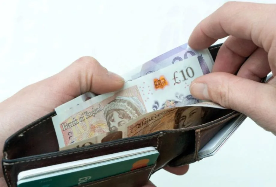 A person opens a brown leather wallet to reveal British banknotes and cards.