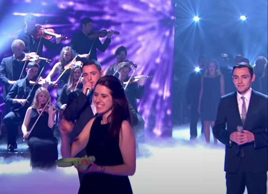 A woman holds a carton of eggs, about to throw one, while a man sings into a microphone with an orchestra behind them.