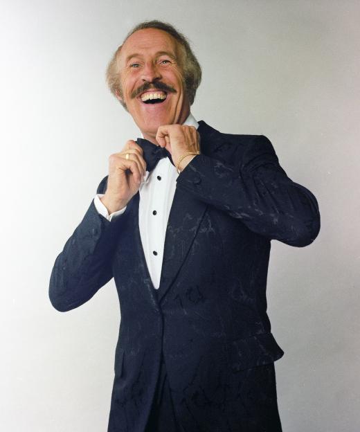 Studio portrait of an older man with a mustache, adjusting his bow tie and laughing while wearing a dark patterned tuxedo jacket over a white ruffled shirt.