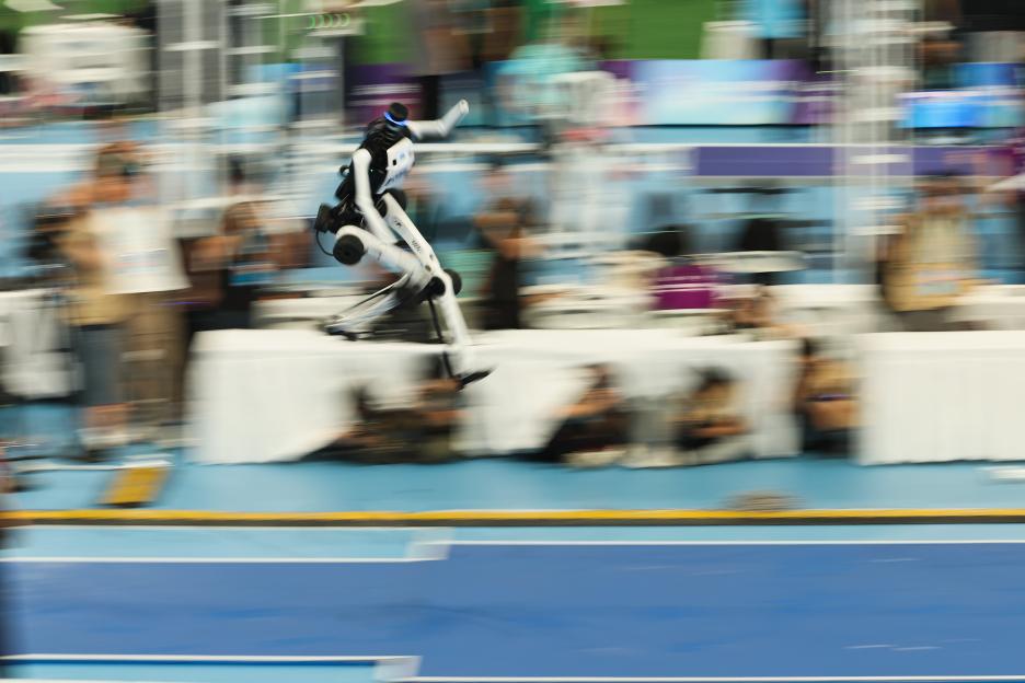 A humanoid robot with black and white body parts in mid-air during a long jump competition.
