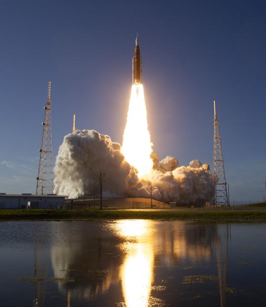 The NASA Space Launch System rocket launching from Kennedy Space Center, reflecting bright orange fire and white smoke on the water.
