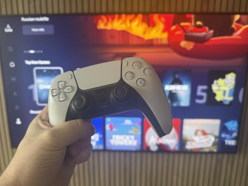 A hand holding a white PlayStation 5 controller in front of a TV screen displaying the console's menu.