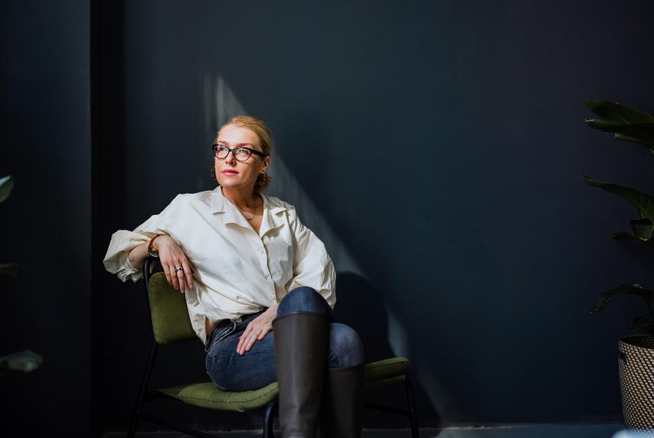 A businesswoman wearing glasses, a white button-down shirt, and jeans with tall brown boots, sits in a chair, looking intently into the distance.