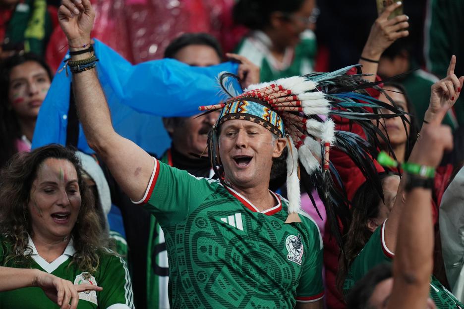 A Mexican fan in a green jersey and feathered headdress cheers with his arm raised in a stadium.