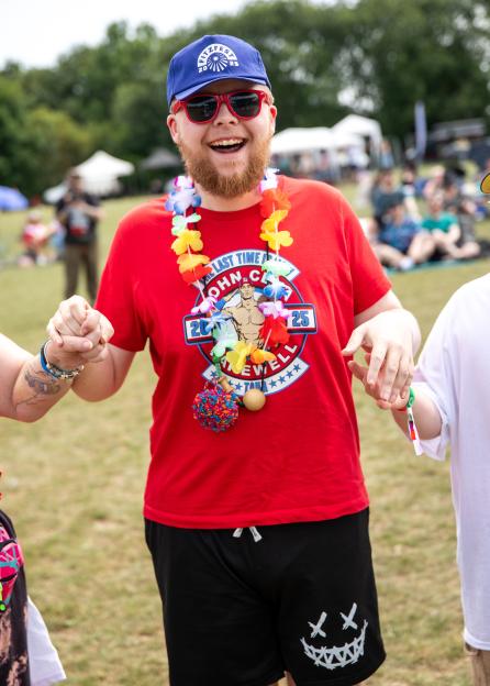 Andre smiling, wearing a red shirt, blue "FITZFEST" cap, red sunglasses, and a colorful lei, holding hands with two people.