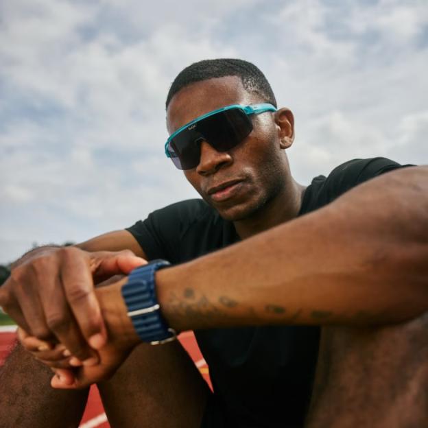 A man wearing turquoise and black athletic sunglasses sits on a running track.