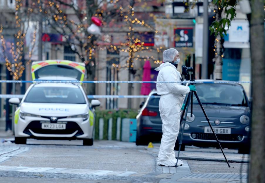 A forensic investigator in a white suit and hairnet takes pictures at a crime scene on Railway Street, Altrincham, marked by police tape and a parked police car.