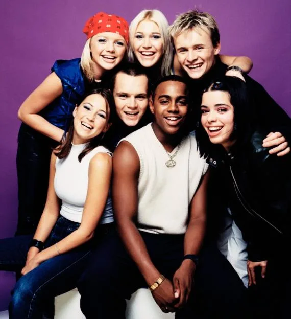 Studio portrait of British pop group S Club 7 posing together in front of a purple background.