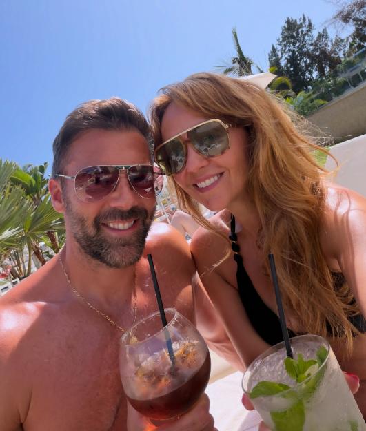 Samia Longchambon and her husband Sylvain on holiday, holding drinks and smiling at the camera.