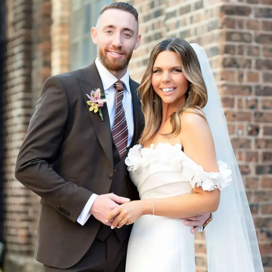 A newlywed couple, a man and a woman, smiling and holding hands, posing in front of a brick building.