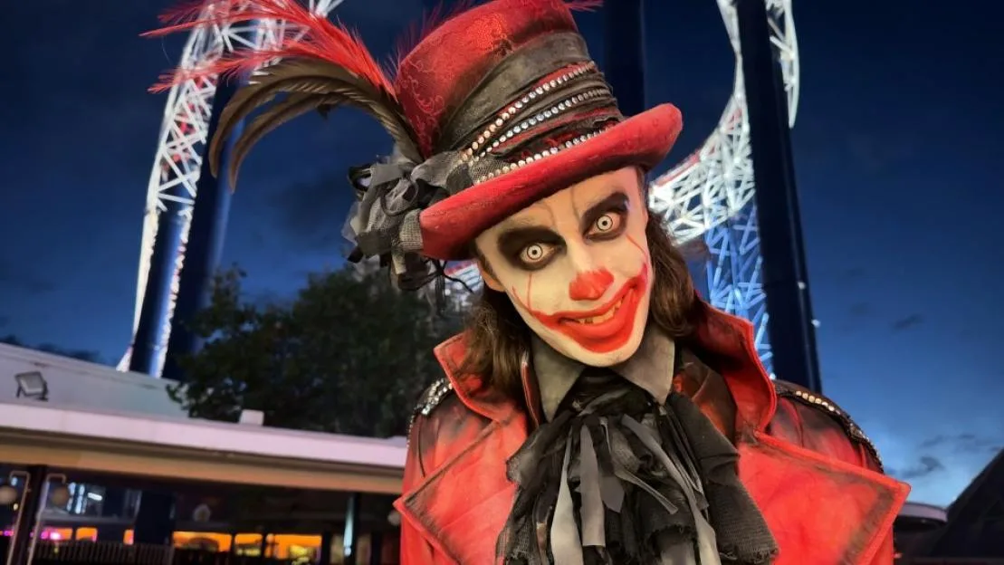 A costumed performer in a red top hat and white and red clown makeup smiles menacingly in front of a lit-up roller coaster at night.