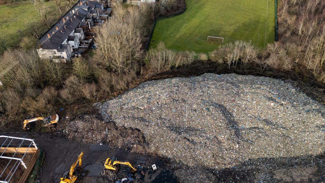 Aerial view of an illegal waste dump in Bickershaw, Wigan, Manchester, with houses and a football field nearby.