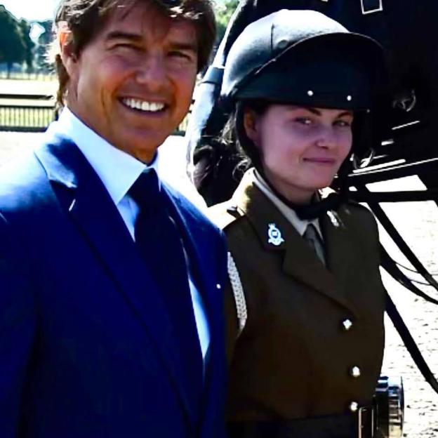 Tom Cruise smiling next to a servicewoman in uniform and equestrian helmet.