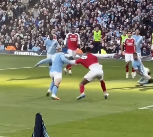 A soccer player in a red jersey attempts to pass another player in a light blue jersey, while a third player in a light blue jersey falls to the ground.