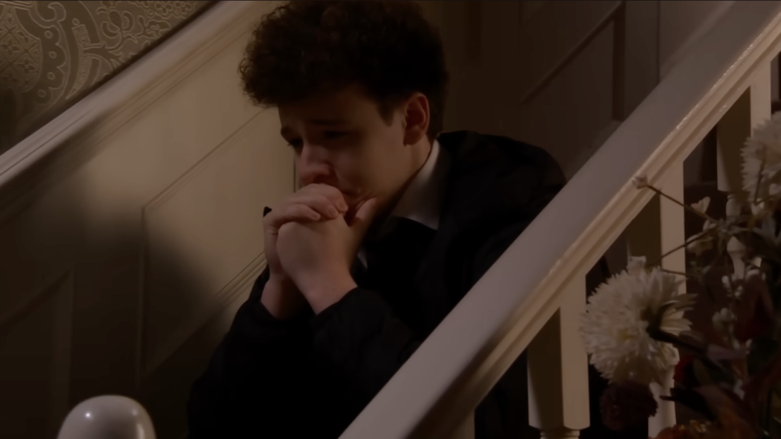 A young man with curly hair sits on a dimly lit staircase, clasping his hands together, looking upset.