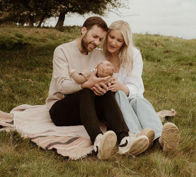 Dan Kettle and Bethan Kettle holding baby Rex while sitting on a blanket in a field.