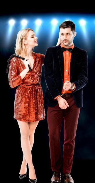A smiling woman in a red sequined dress holding a whip stands beside a man in a suit with his hands cuffed.