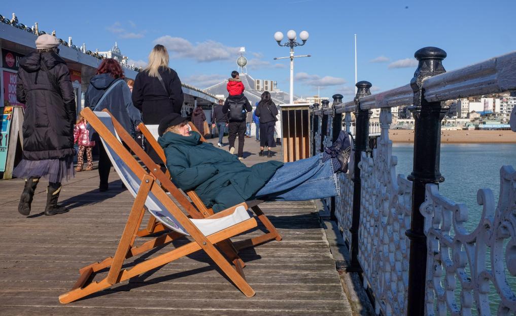 A person in a heavy winter coat and jeans relaxes in a deckchair on Brighton Palace Pier, facing the ocean.