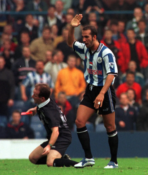 A soccer player in a blue and white striped jersey argues with a referee who is on his knees on the field.