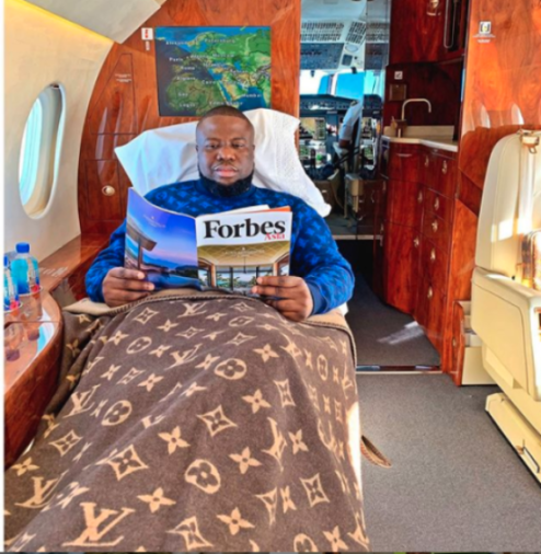 Ramon Olorunwa Abbas, also known as 'Hushpuppi', sitting on a private jet, covered with a Louis Vuitton blanket, and reading a Forbes Asia magazine.
