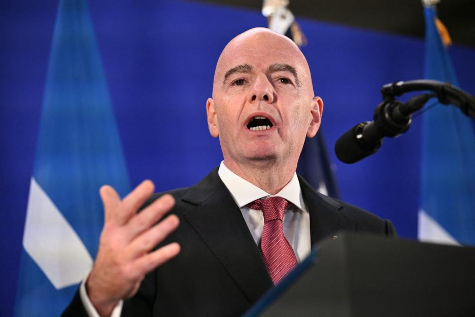 A man with a bald head and red tie speaks into a microphone, gesturing with his left hand.